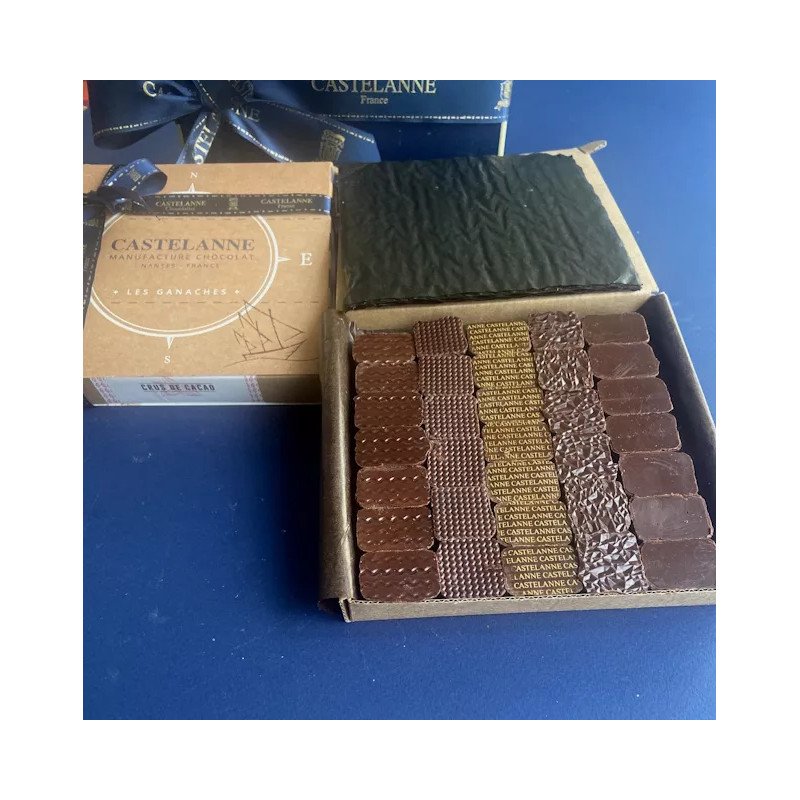 Boîte ouverte de chocolats Castelanne assortis en plusieurs tailles et textures, alignés dans un rangées sur fond bleu.