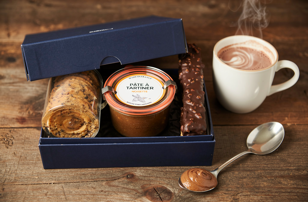 Coffret gourmand avec pâte à tartiner noisette, cookies et bâton chocolaté posé près d'un chocolat chaud maison.