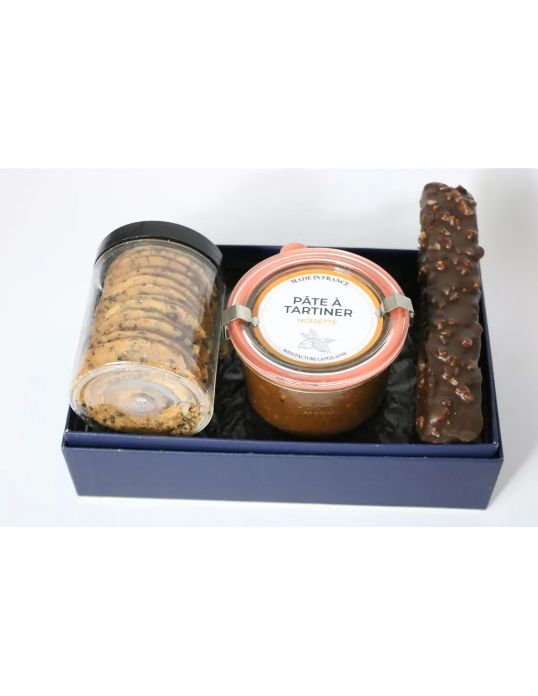Assortiment gourmet: biscuit roulé noir, pâte à tartiner noisette en pot, et barres chocolatées sur un socle bleu.