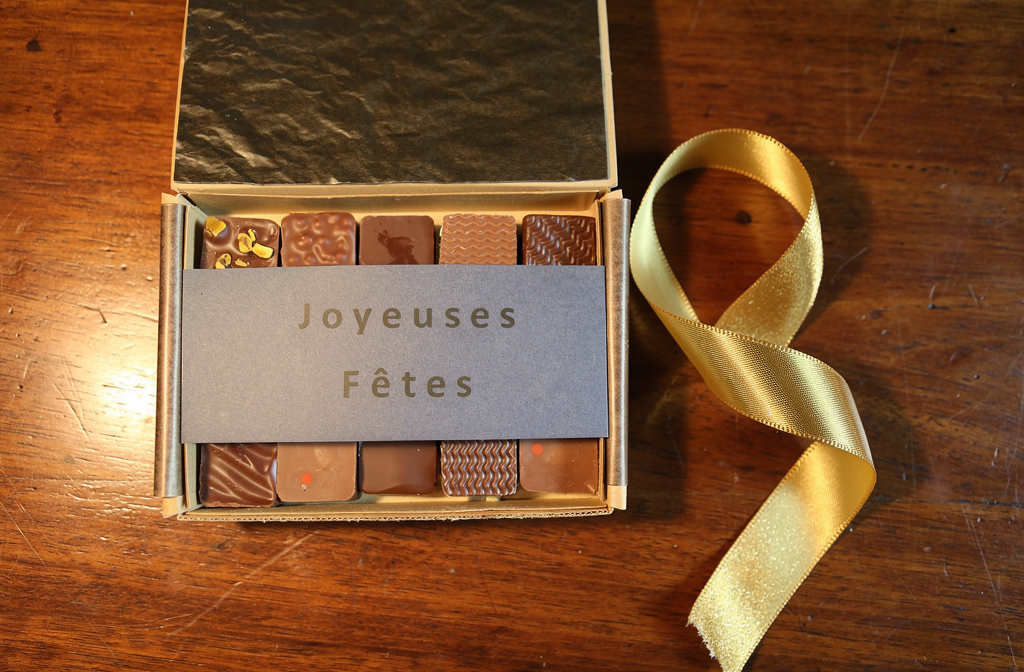 Boîte de chocolats artisanaux Castelanne, ouvert avec une carte "Joyeuses Fêtes" et ruban doré sur bois.
