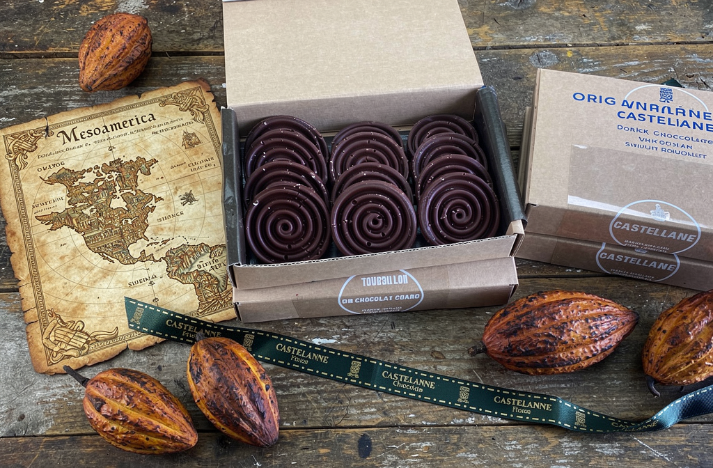 Boîte de tourbillons de chocolat noir Castelanne posée sur une table en bois avec des fèves de cacao et une carte ancienne.