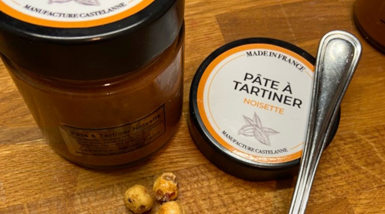 Pâte à tartiner noisette Castelanne en pot ouvert, cuillère argentée et billes de noisettes à côté sur bois. Pâte à tartiner gourmande Castelanne noisette en pot fermé avec cuillère et noisettes autour sur plan de bois.